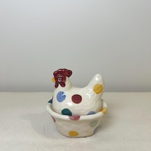 May include: A ceramic chicken-shaped container with a red comb and yellow beak. The white body is decorated with colourful polka dots. The chicken sits atop a matching bowl, also dotted.