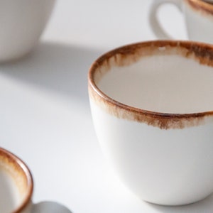 May include: A white ceramic coffee mug with a brown rim and a brown stain around the top edge. The mug is empty and sitting on a white surface.