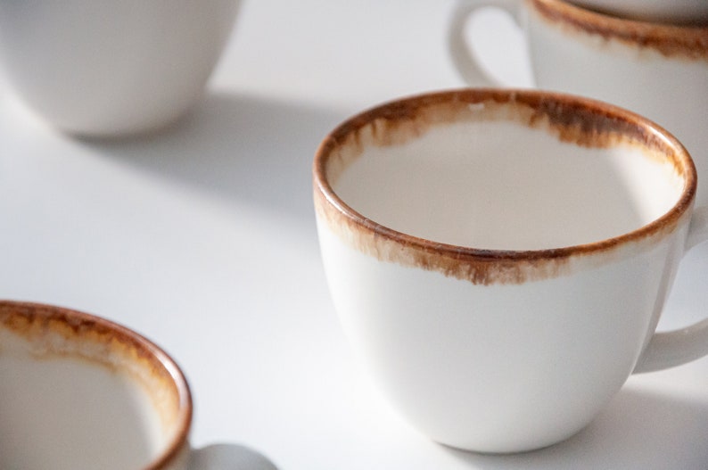 May include: A white ceramic coffee mug with a brown rim and a brown stain around the top edge. The mug is empty and sitting on a white surface.