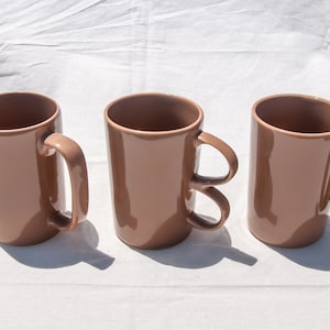 Peut inclure: Trois tasses en céramique marron avec des styles de poignées différents. Les tasses sont toutes de la même taille et de la même forme.