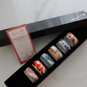 Puede incluir: Un juego de seis servilleteros de cerámica Clarice Cliff de la colección Napkin Ring, cada uno con un diseño colorido único. Los anillos se muestran en una caja negra con un certificado.