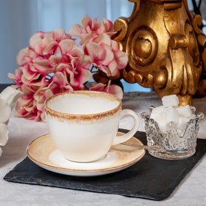 May include: A white teacup and saucer with a brown rim, a white teapot with a rose design, and a glass sugar bowl in the shape of a crown filled with white sugar cubes. The tea set is on a black slate surface with pink flowers in the background.