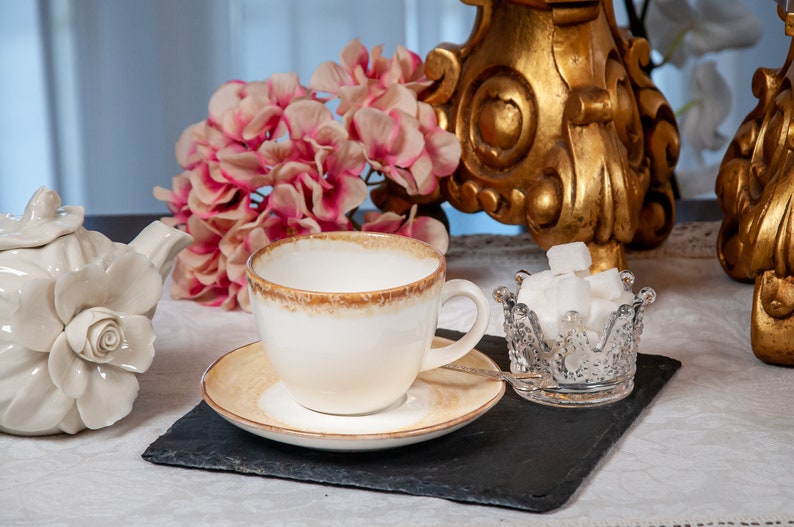 May include: A white teacup and saucer with a brown rim, a white teapot with a rose design, and a glass sugar bowl in the shape of a crown filled with white sugar cubes. The tea set is on a black slate surface with pink flowers in the background.