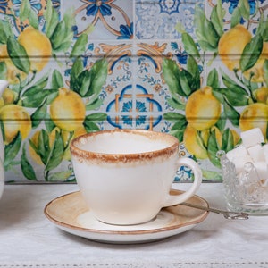 May include: A white teacup and saucer with a brown rim, a white teapot, and a glass sugar bowl filled with white sugar cubes. The background is a tiled wall with a lemon pattern.