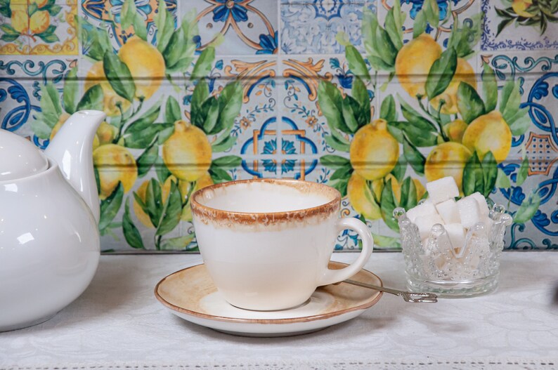 May include: A white teacup and saucer with a brown rim, a white teapot, and a glass sugar bowl filled with white sugar cubes. The background is a tiled wall with a lemon pattern.