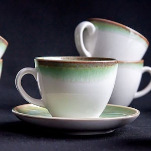 May include: White ceramic teacups and saucers with a green and brown rim. The teacups have a curved handle and are stacked in a group. The saucers are round and match the teacups. The background is dark.
