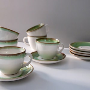 Może przedstawiać: Zestaw białych ceramicznych filiżanek i spodków z zielono-brązową obwódką. Filiżanki są ułożone w stos i ustawione na białej powierzchni. Spodki są ułożone osobno. Idealne do serwowania herbaty lub kawy.