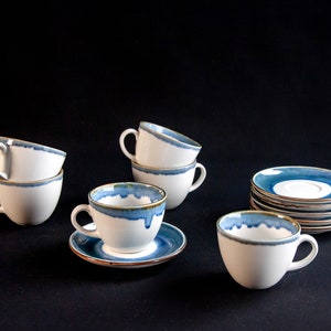May include: Set of six white ceramic teacups and saucers with blue and gold accents. The cups and saucers have a unique, hand-painted design with a blue and gold glaze.