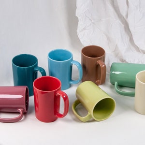Peut inclure: Huit tasses en céramique de différentes couleurs, dont le rouge, le bleu, le vert, le marron, le rose et le jaune. Les tasses sont disposées sur une surface blanche.