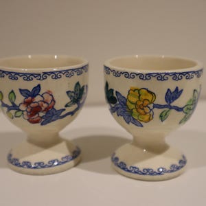 Puede incluir: Dos hueveras color crema con diseños florales. Cada taza presenta una base de pedestal y un borde con un patrón de desplazamiento azul. Los patrones florales incluyen acentos rojos, amarillos y azules.