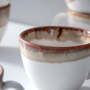 May include: A white ceramic coffee mug with a brown and beige glaze. The mug has a rounded shape and a handle.