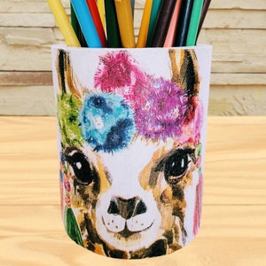 May include: A colorful llama pencil holder with a white background and a floral design around the head. The holder is filled with colored pencils.