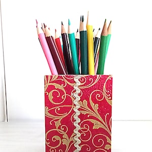 May include: A red pencil holder with a gold floral pattern and a white ribbon. The holder is filled with coloured pencils.