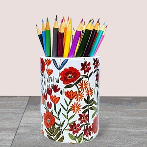 May include: A white ceramic pencil holder with a floral pattern featuring red, orange, and yellow flowers. The holder is filled with a variety of colored pencils.
