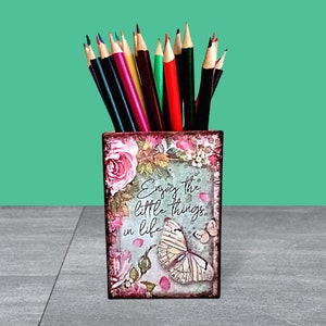 May include: A floral patterned pencil holder with the text "Enjoy the little things in life" and a butterfly design. The holder is filled with colored pencils.