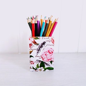 May include: A white ceramic pencil holder with a floral pattern and the words "Trend, unique, flowers, beauty, elegant" printed on it. The holder is filled with colored pencils.