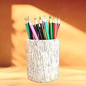 May include: A collection of colored pencils in a white, textured pencil holder. The pencils are various colors, including pink, green, and blue. The pencil holder has a bark-like texture.