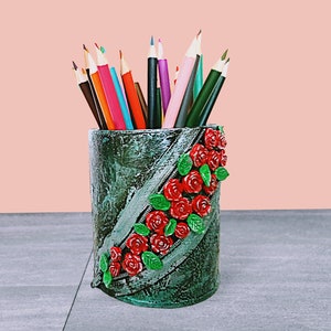 May include: A green and gray ceramic pencil holder with a zipper design and red roses. The holder is filled with colored pencils.