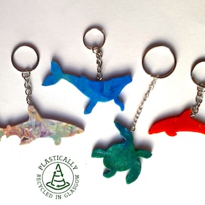 Puede incluir: Cinco llaveros de animales de plástico de colores, incluyendo una ballena, un tiburón, una tortuga y un delfín. Los llaveros están hechos de plástico reciclado y llevan el logotipo "Plastically Recycled in Glasgow".