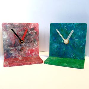 Puede incluir: Dos relojes de escritorio cuadrados. Uno es rojo, rosa y gris con manecillas negras y rojas. El otro es verde y azul con manecillas blancas. Ambos relojes tienen un aspecto veteado y una base pequeña.