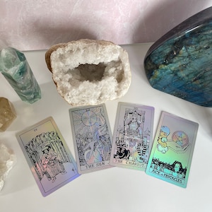 May include: A spread of four tarot cards on a white surface with crystals and stones. The cards are The Sun, The Chariot, Ace of Pentacles, and IX. The cards are iridescent and have black and white illustrations.