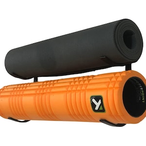 May include: Black metal wall mount with two storage shelves. A black yoga mat is on the top shelf and an orange foam roller is on the bottom shelf. The foam roller has a black and yellow "Y" logo and the text "TriggerPoint" on the side.