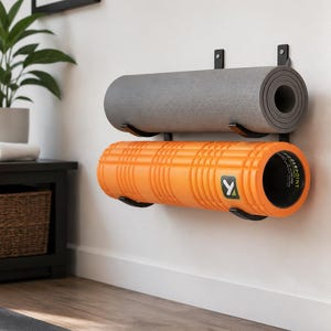 May include: A black metal wall-mounted rack holding two cylindrical exercise items. The top item is a solid gray yoga mat, and the bottom item is a bright orange textured foam roller with a black interior and a TriggerPoint brand logo.