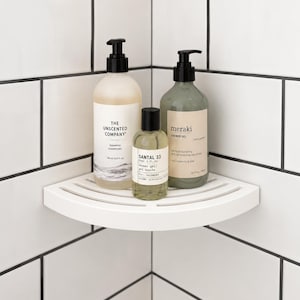 May include: A white corner shower shelf holding three bottles: a large beige bottle labeled The Unscented Company Shampoo, a smaller clear bottle labeled Santal 33 Shower Gel, and a large sage green bottle labeled Meraki Shower Gel with black pumps.