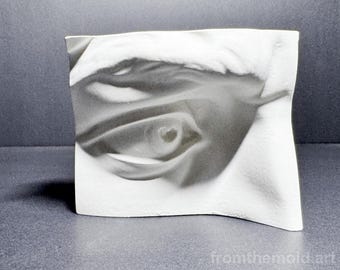 May include: A rectangular white sculpture with a wavy, undulating surface featuring a grayscale photographic print of a human eye. The image shows the iris, pupil, and surrounding eyelid details. The text fromthemold.art is printed in the corner.