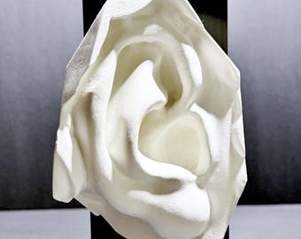 May include: A white, three-dimensional sculptural wall hanging shaped like a human ear, featuring a textured matte surface. The piece is mounted on a vertical black rectangular panel and includes a small silver metal loop at the top for hanging.