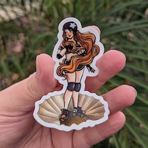 May include: A sticker featuring a cartoon illustration of a roller skater with long orange hair, a helmet, and knee pads, standing in a seashell. The skater is wearing a black top and shorts, and roller skates. The sticker is held in a hand.