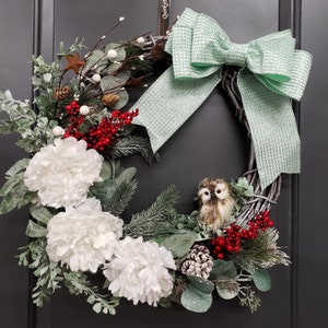 May include: A winter-themed wreath with white flowers, red berries, and a small owl figurine. A green and white bow is tied to the wreath.