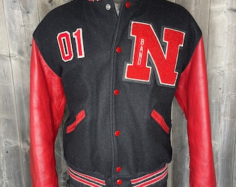 Op de afbeelding: Zwart varsity jack met rode leren mouwen. Het jack heeft een rode vilt 'N' met 'BAND' verticaal geschreven op de linker borst. Het nummer '01' staat op de rechter borst. Het jack heeft een witte en rode gestreepte rand rond de kraag, manchetten en onderkant.