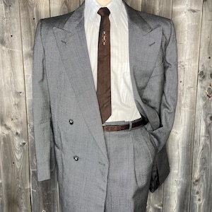 Puede incluir: Un traje gris de doble botonadura con camisa blanca y corbata marrón. El traje tiene un diseño clásico con solapa de muesca y pantalón con pliegues. Un cinturón de cuero marrón completa el look. El traje se exhibe en un maniquí.