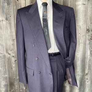 May include: A dark purple double-breasted suit jacket with wide lapels and three visible buttons, paired with a white collared shirt and a grey patterned necktie featuring a silver tie bar with a chain, worn over matching pleated trousers.
