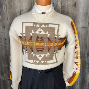 May include: A cream-colored sweater with a brown, orange, and yellow geometric pattern. The sweater has a crew neck and long sleeves.