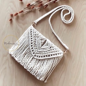 Könnte beinhalten: Eine weiße Makramee-Tasche mit Fransenrand und langem Trageriemen. Die Tasche ist in einer Knotentechnik gefertigt und hat ein geometrisches Muster.