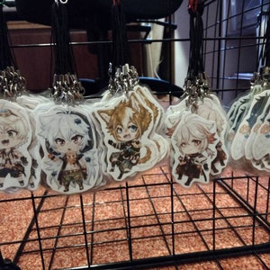 Puede incluir: Una colección de ocho llaveros de personajes de estilo anime, cada uno con un personaje diferente con diseños y colores únicos. Los llaveros se muestran en una rejilla de alambre negro.