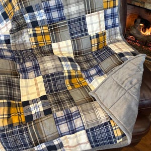 May include: A patchwork quilt with a variety of plaid patterns in shades of blue, gray, yellow, and white. The quilt is draped over a brown leather chair, with a gray, plush backing visible. The quilt's design is a mix of squares.
