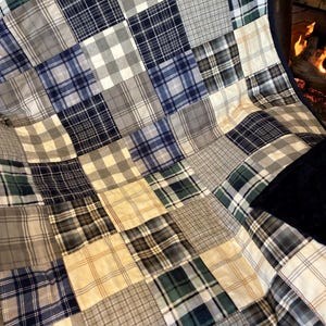 May include: A patchwork quilt with a plaid pattern in shades of blue, green, gray, and cream. The quilt is made of squares sewn together, creating a textured surface. The quilt is draped over a dark surface.