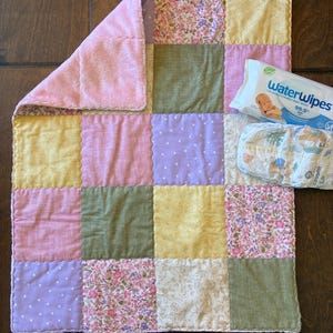May include: A patchwork baby blanket with squares of pink, yellow, green, and floral fabrics. A package of WaterWipes baby wipes and a diaper are next to the blanket. The blanket has a soft pink plush border.