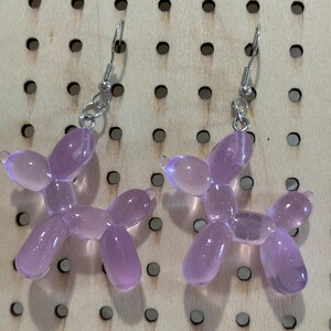 Puede incluir: Un par de pendientes de perro globo de color lavanda. Los pendientes están hechos de plástico translúcido y tienen un gancho plateado. Los pendientes se exhiben en un tablero de clavijas de madera.