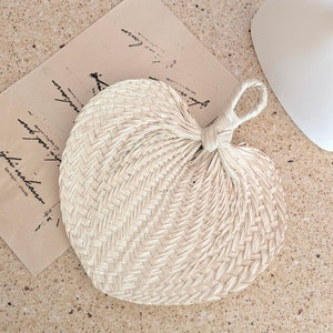 May include: A heart-shaped hand fan made of woven natural fibers. The fan has a looped handle and a tightly woven pattern. The fan is lying on a light-colored surface next to a piece of paper with script.