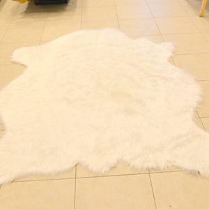 May include: A large, off-white faux fur rug with an irregular shape, resembling an animal hide. The rug is placed on a tiled floor, and the fur appears soft and plush. The rug's dimensions are approximately 60 inches long and 40 inches wide.