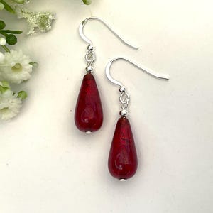 May include: A pair of drop earrings featuring smooth, teardrop-shaped red glass beads suspended from silver-toned metal hooks. Each bead is accented with a small silver-toned spacer bead at the top, connecting the glass to the curved ear wire.