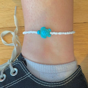 May include: Ankle bracelet featuring a turquoise turtle charm. The bracelet is made of small white beads and is worn on an ankle above a black canvas shoe. The turtle charm is centered on the bracelet.