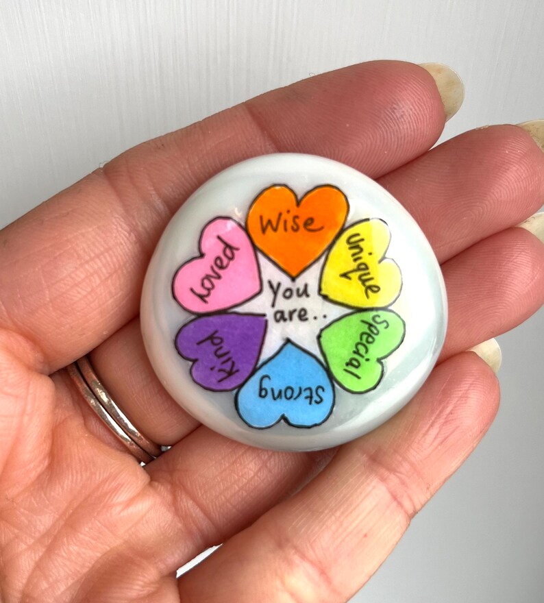 May include: A white, round, decorative stone with a colorful heart design. Each heart is a different color and contains a positive word: Loved, Kind, Strong, Special, Unique, and Wise. The words "You are.." are in the center.