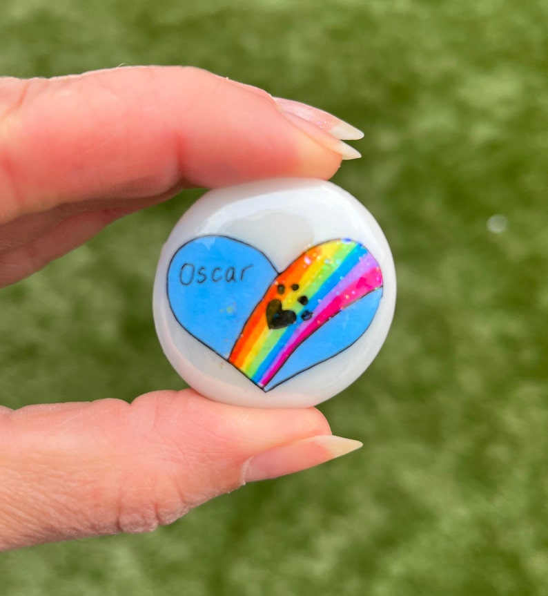 May include: A white heart-shaped stone with a blue heart painted on it. The blue heart has the name "Oscar" written on it and a rainbow with paw prints painted on it.