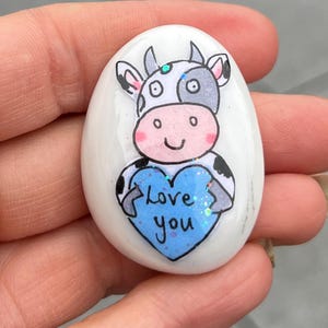 May include: A white painted rock with a cartoon cow holding a blue heart that says "love you" in white letters.