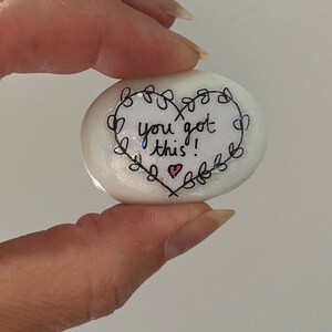 May include: White heart-shaped stone with black ink lettering that reads "You got this!" surrounded by a black floral design. A small red heart is drawn at the bottom of the stone.